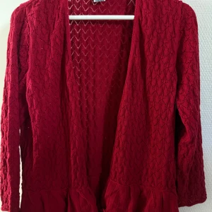 Röd stickad kofta med volangkant - Snygg röd stickad kofta med lång ärm och öppen front. Koftan har ett genomgående hålmönster och dekorativ volangkant nedtill som ger en extra touch. Perfekt för dig som vill ha något både gulligt och trendigt i garderoben.