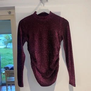 Glittrig vinröd/lila topp från Gina Tricot - Säljer en superfin vinröd glittrig topp från Gina Tricot. Långa ärmar och en lite högre hals. Den sitter ganska tajt runt armarna.
