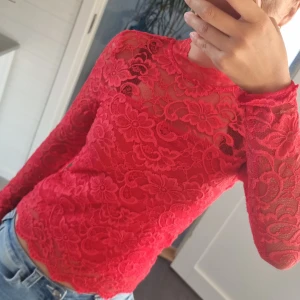 Röd spetsblus med öppen rygg - Säljer en röd långärmad blus i spets med blommigt mönster. Toppen har en snygg öppen rygg med knapp i nacken och vågig kant vid ärmslut och nederkant. Perfekt för dig som vill sticka ut med en feminin och trendig look.