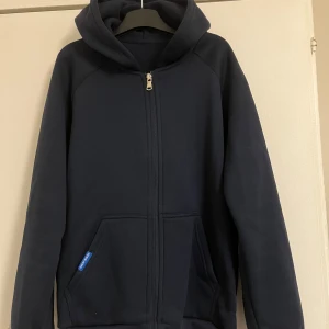 Mörkblå hoodie med dragkedja - Mörkblå hoodie från Grizzly med hel dragkedja, huva och två fickor framtill. Tröjan är i mjuk bomullsmix och har en klassisk, enkel design som passar perfekt till chill dagar. Snygg och basic stil med normal passform.