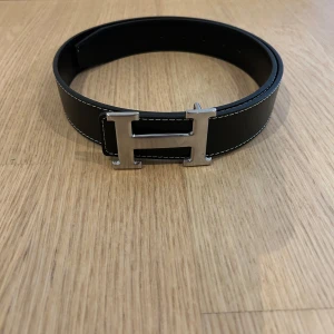 Svart H skärp med H-spänne - Säljer ett svart skärp med ikoniskt H-spänne i silverfärgad metall. Bältet är gjort i grynigt skinn med vita kontrastsömmar och har en bred, stilren design som verkligen sticker ut. Perfekt accessoar för att lyfta vilken outfit som helst.
