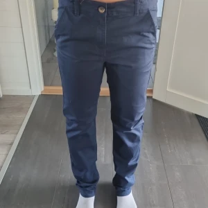Mörkblå chinos från Kappahl, stl 170 - Snygga mörkblå chinos från Kappahl i storlek 170. Byxorna har klassisk passform med raka ben, sidfickor och bakficka med liten etikett. Tillverkade i mjuk bomull som känns skön hela dagen. Perfekta för en clean och stilren look.