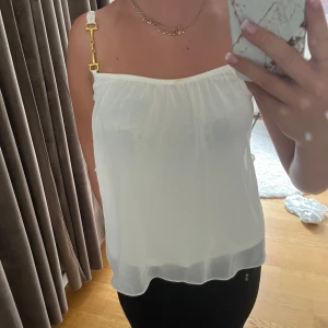 Vit linnetopp från Ida Sjöstedt - Superfin vit linnetopp från Ida Sjöstedt med tunna axelband och snygga guldfärgade metalldetaljer. Toppen är lätt transparent och har en luftig, flowy passform som känns riktigt somrig. Perfekt att styla med jeans eller kjol för en fräsch look. Linnet har två lager så den är inte så genomskinlig (på bilden har jag en svart bh)