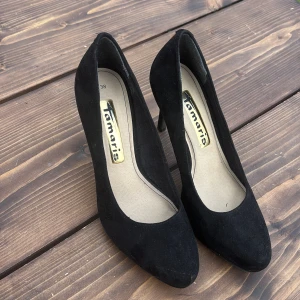 Svarta pumps från Tamaris i mocka - Stilrena svarta pumps från Tamaris i mjuk mocka med klassisk rund tå och hög klack. Perfekta för dig som vill ha en tidlös och elegant sko till din outfit. 