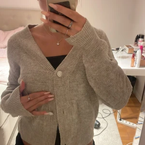 Beige ribbad kofta med glittriga knappar - Supermysig beige kofta med ribbad struktur och glittriga knappar framtill. Koftan har en djup v-ringning och långa ärmar med lite oversized känsla. Perfekt att slänga över ett linne eller t-shirt för en chill look.