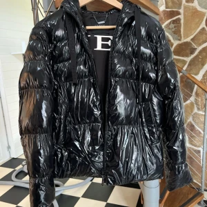 Svart pufferjacka från Guess - Snygg svart pufferjacka från Guess med glansig finish och huva. Ca använt 4 gånger . Den är vintage går ej att få dag på. Köpt för 2500kr Klicka ej på köp 