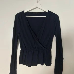 Marinblå omlottopp från Bik Bok - Snygg marinblå omlottopp från Bik Bok med långa ärmar och v-ringning. Toppen har en markerad midja och volangkant nedtill som ger en feminin siluett. Perfekt att styla med jeans eller kjol för en trendig look.