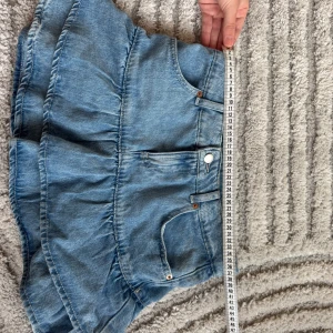 Blå volangkjol i jeans - Supersöt kort jeanskjol i ljusblå denim med volanger och klassiska fickor fram. Kjolen har en feminin och lekfull look med markerad midja och knappstängning. Perfekt för dig som gillar en trendig och avslappnad stil.