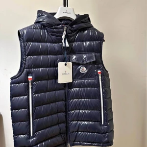 Mörkblå dunväst från Moncler - Snygg mörkblå dunväst från Moncler med glansig finish, huva och raka quiltade sömmar. Västen har två dragkedjefickor fram, en bröstficka med lock och Moncler-logga. Perfekt för lager-på-lager och streetstyle.