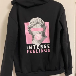 Svart hoodie med tryck och huva - Svart hoodie med stor ficka framtill och snörning i huvan. Baksidan har ett coolt tryck med antik staty, rosa detaljer och texten 'INTENSE FEELINGS'. Perfekt för dig som gillar streetstyle och unika prints.