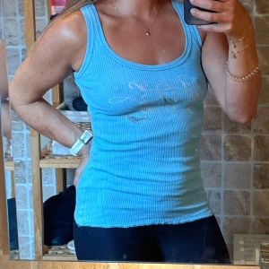 Ljusblå ribbad linnetopp - Säljer en ljusblå ribbad linnetopp i storlek M. Toppen har ett snyggt tryck med glansiga bokstäver framtill och är ärmlös med rund halsringning. Perfekt för en chill och sportig look.