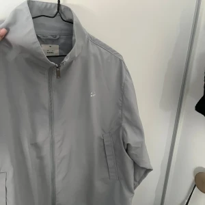 Ljusgrå vindjacka från A DSGN - Snygg ljusgrå vindjacka från A DSGN med minimalistisk design. Jackan har hög krage, dragkedja framtill och två fickor på sidorna. Perfekt för dig som gillar stilrena plagg med sportig vibe. Materialet är lätt och känns smidigt att bära. Slutsåld på A dsgn hemsida! Helt oanvänd. Har en i svart som jag använder istället 
