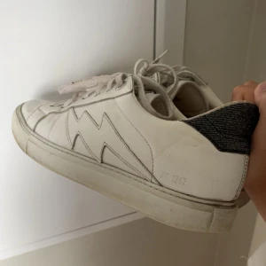 Vit sneaker Zadig & Voltaire blixtdetalj - Snygga vita sneakers från Zadig & Voltaire med ikonisk blixtdetalj på sidan och svart glittrig häl. Skorna har snörning och är tillverkade i skinn med platt sula och rund tå. Köpta på Plick men har inte kommit till så mycket användning