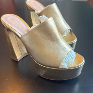 Guldiga platåklackar från Gianvito Rossi Milano - Statementklackar i metallicguld från Gianvito Rossi med bred rem över foten, öppen tå och kraftig platåsula. Skorna har hög, skulptural klack och är tillverkade i skinn med lyxig finish. Perfekta för dig som vill sticka ut med en glamorös look.