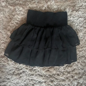 Svart volangkjol med smockmidja - Supersöt svart kortkjol med bred smock i midjan och dubbla volanger. Kjolen har en luftig och flowy känsla, perfekt för att skapa en cool och trendig look. Passar dig som gillar att sticka ut med en enkel men snygg stil. Använd fåtal gånger🤍