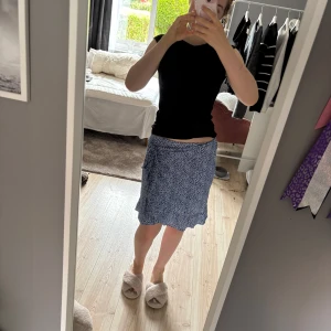 Blå omlottkjol med småblommigt mönster - Supersöt blå omlottkjol med vitt småblommigt mönster. Kjolen har knyt i sidan och en lätt vågig nederkant som ger en avslappnad vibe. Perfekt för varma dagar och enkel att matcha med en basic topp.