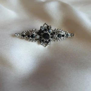 Vintageinspirerad silverfärgad brosch - En detaljrik brosch i silverfärg med gnistrande svarta och klara stenar. Den har en avlång, spetsig form med vackra ornament och stenar i olika storlekar.