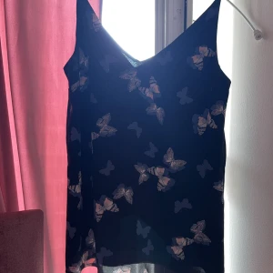 Svart linnetopp med fjärilar från Oasis - Svart linnetopp från Oasis med v-ringning och tunna axelband. Toppen har ett snyggt fjärilsmönster i lila och rosa toner som ger en cool och lekfull vibe. Perfekt att styla med jeans eller kjol för en trendig look.