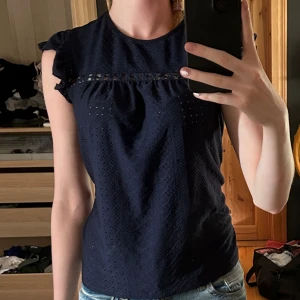 Mörkblå blus med volangärmar - Supersöt mörkblå blus med broderade detaljer och volangärmar. Toppen har små hålmönster och en fin spetskant över bröstet. Perfekt till jeans eller kjol för en chill och stilren look.