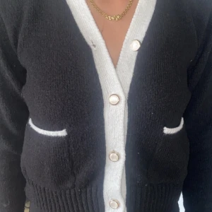 Svart kofta med vita detaljer och knappar - Säljer en svart kofta med vita kanter och snygga pärlknappar. Koftan har två små fickor framtill med vita detaljer och ribbad nederkant. Perfekt för dig som gillar klassisk men trendig stil. Långärmad och stickad för extra mysfaktor.