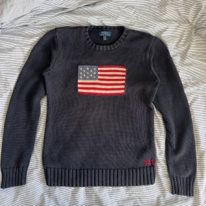  Polo Ralph Lauren amerikan flag  - Snygg mörkblå stickad tröja från Polo Ralph Lauren med amerikansk flagga på bröstet och RL-broderi i rött vid nederkanten. Tröjan har rund halsringning, ribbade muddar och lång ärm. Storlek 18-20 vilket sitter ungefär som small Perfekt för dig som gillar klassisk preppy stil.