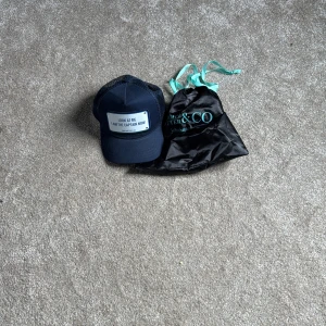 Marinblå keps John Hatter & Co - Snygg marinblå keps från John Hatter & Co med justerbar baksida i mesh och en metallskylt framtill med texten 'LOOK AT ME I AM THE CAPTAIN NOW'. Kepsen kommer med originalpåse och har en klassisk trucker-modell.