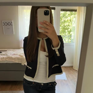 Mörkblå kofta med vita detaljer - Snygg mörkblå kofta med vita kantdetaljer och dekorativa knappar vid ärmsluten. Koftan är kortare i modellen och har två små fickor framtill. Perfekt för en chic och stilren look.