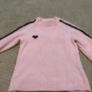 Rosa stickad tröja med hjärta från Monki - Supergullig ljusrosa stickad tröja från Monki med mörkbruna ränder längs ärmarna och ett mörkbrunt hjärta på bröstet. Tröjan har ribbade muddar och rund hals. Perfekt för dig som gillar söta detaljer och vill ha en mysig vibe.