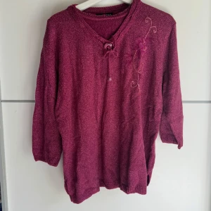 Mörklila stickad tröja från Domitex - Mysig mörklila stickad tröja från Domitex med trekvartsärm och v-ringning. Tröjan har en snygg snörning vid halsen och broderad blomdetalj på ena axeln. Perfekt för dig som gillar unika detaljer och en chill vibe.