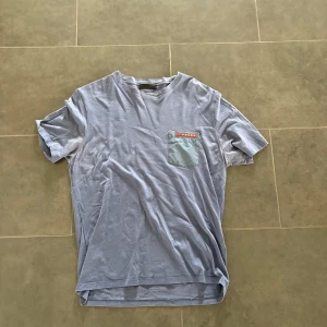 Ljusblå t-shirt från Prada - Snygg ljusblå t-shirt från Prada med en liten bröstficka i matchande färg och röd logodetalj. Klassisk passform och rund halsringning, perfekt för en clean och stilren look.