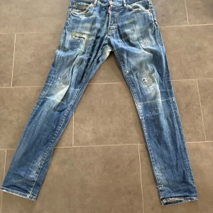 Blå slitna jeans från Diesel - Säljer ett par blå jeans från Diesel med coolt sliten look, färgstänk och lagade detaljer. Bakfickan har en stor broderad D. Jeansen har smal passform och klassisk femficksdesign. Perfekta för dig som gillar en edgy streetstil.