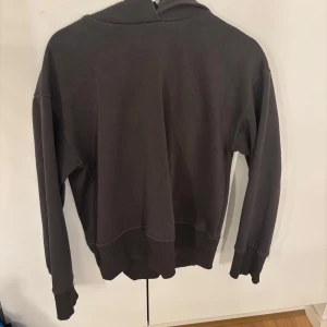 Mörkgrå hoodie med huva - Enkel och stilren mörkgrå hoodie med stor huva och långa ärmar. Tröjan har breda muddar vid ärmslut och nederkant för en skön passform. Perfekt för chill dagar eller när du vill ha en avslappnad look.