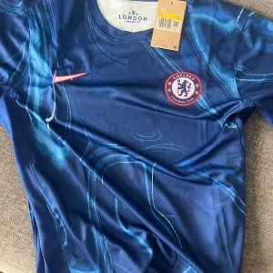 Chelsea FC blå matchtröja Nike S - Chelsea FCs officiella matchtröja från Nike i blått med unikt mönster och klubbmärke på bröstet. Tröjan har korta ärmar, rund hals och är tillverkad i ett lätt, ventilerande funktionsmaterial. Perfekt för dig som vill visa vilket lag du håller på.