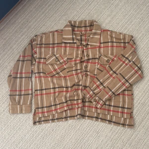 Beige rutig overshirt i bomull - Snygg beige overshirt i 100% bomull med rutigt mönster i svart, vitt och rött. Skjortan har två bröstfickor med lock och knappar, klassisk krage och långa ärmar. Perfekt att slänga över en t-shirt för en chill look.