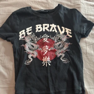 Svart ribbad t-shirt med draktryck - Cool svart ribbad t-shirt från New Yorker i storlek XS. Framsidan har ett stort tryck med två drakar, röd cirkel och texten 'BE BRAVE' samt asiatiska tecken. T-shirten är kortärmad och har en tight passform. Använd fåtal gånger så den är som ny. 