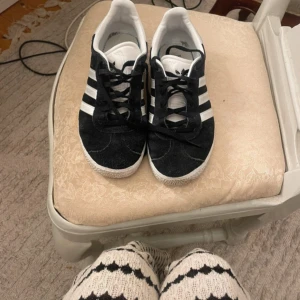 Svarta Adidas Gazelle sneakers mocka - Klassiska Adidas Gazelle sneakers i svart mocka med vita detaljer och de ikoniska tre ränderna på sidorna. Låg modell med snörning och vit sula med mönster undertill. Perfekta för en clean och sportig look.