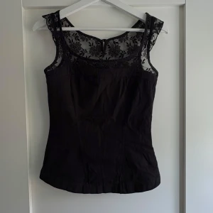 Svart blommig spetsblus - Supersnygg svart blus med blommig spets upptill och ärmlös design. Toppen har rundad halsringning och feminina detaljer i spetsen som ger en elegant vibe. Perfekt att styla med jeans eller kjol för en trendig look.