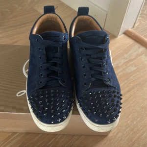 Blå Christian Louboutin sneakers med nitar - Snygga marinblå sneakers från Christian Louboutin i mocka med svarta nitar på tån och klassisk röd sula. Skorna har snörning, rund tå och vita gummisulor. Kommer med originalkartong, dustbag och äkthetskort. Perfekta för dig som vill sticka ut med en edgy vibe. Saknar 5 nitar 