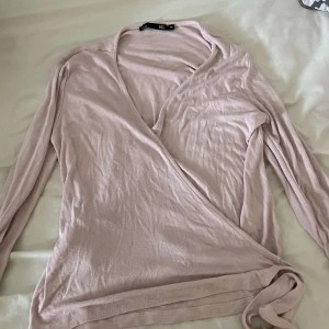 Ljusrosa omlottopp från 157, stl M - Supersöt ljusrosa omlottopp från 157 i mjuk bomull. Långärmad med knyt i sidan och v-ringad hals. Perfekt för dig som gillar en enkel men ändå feminin stil. Sitter snyggt och är lätt att matcha med jeans eller kjol.