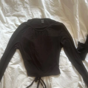 Svart långärmad topp med knyt  - Svart långärmad topp från SHEIN med rund halsringning och knytband i ryggen . Modellen är croppad och har en enkel, stilren look som passar till många outfits. Tunn och mjuk i materialet, med en öppning bak på ryggen 