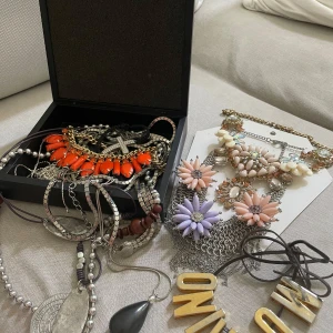Mix av statement- och kedjehalsband - Samling med flera halsband: två statement-halsband med stora blommor i pastellrosa, lila och vitt, dekorerade med gnistrande stenar och kedjor i silver och guld. Flera långa halsband i silverfärg, med hängen i svart, metall och trädetaljer. Perfekt för dig som gillar att mixa stilar och sticka ut.