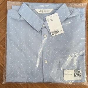 Ljusblå mönstrad skjorta från H&M - Snygg ljusblå skjorta från H&M med små vita mönster och klassisk krage. Skjortan har knappar framtill och är tillverkad i bomull som är lätt att stryka. Perfekt för dig som vill ha en stilren look med lite extra detalj. köptes för 173 säljer för 49kr först till kvarn i orgnal paket.