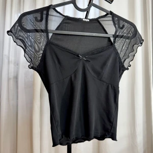 Svart mesh topp med rosett - Snygg svart topp med mesh-ärmar och liten rosett framtill. Toppen har v-ringning och transparenta, vågiga ärmar som ger en cool och trendig look. Perfekt att styla med jeans eller kjol för en edgy vibe.