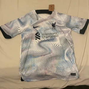 Liverpool Nike bortatröja L - Liverpool FC bortatröja från Nike i storlek L. Tröjan har ett unikt psykedeliskt mönster i vitt med svarta, blå och lila vågiga linjer. Klassisk rund hals, korta ärmar och svart Nike-logga samt LFC-märke på bröstet. Tillverkad i lätt och ventilerande polyester.