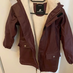 Vinröd regnjacka från Didriksons - Snygg vinröd regnjacka från Didriksons med mysigt vitt fleecefoder. Jackan har huva, dragkedja och stora fickor med tryckknappar. Nypris är ca 600 kr.