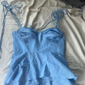 Ljusblå korsett-topp med volang - Supersöt ljusblå korsett-topp med tunna knytbara axelband och volang nertill. Toppen har formade kupor och smock i ryggen för extra komfort. Perfekt för dig som vill ha en feminin och trendig look med en touch av sommar.