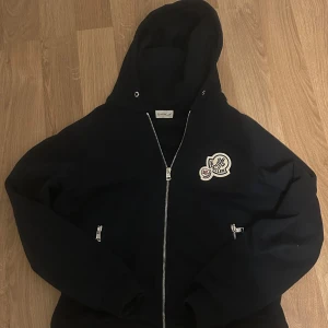 Svart hoodie från Moncler - Snygg svart hoodie från Moncler med dragkedja och broderad logga på bröstet. Perfekt för en stilren och bekväm look. Den har en justerbar huva och praktiska fickor med dragkedjor. Funkar för L och M. Moncler tagg ingår och Nfc funkar (kan lösa bild), Hoodie snören finns kvar med. Kom dm för eventuella frågor. Priset är ej hugget i sten🙂