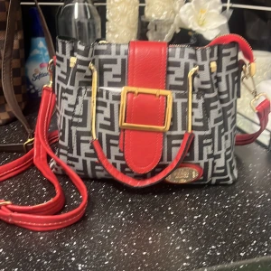 Fendi-inspirerad axelväska med rött läder - Snygg axelväska med Fendi-inspirerat svart och grått mönster, detaljer i rött läder och guldiga metalldetaljer. Väskan har en bred röd axelrem, stor guldfärgad spänne och dragkedja upptill. Perfekt för dig som vill sticka ut med en trendig accessoar.