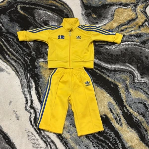 Gula Adidas byxor med blå ränder - Snygga gula Adidas byxor för små barn med klassiska blå ränder längs sidorna och Adidas-logga framtill. Byxorna har resår i midjan för skön passform och är gjorda i mjukt material som passar perfekt för lek och rörelse.