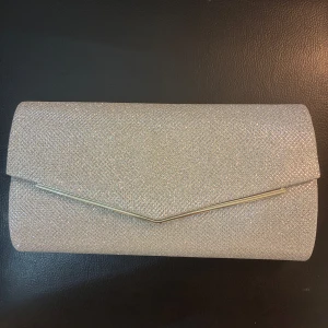 Glittrig silver kuvertväska med kedja - Snygg kuvertväska i glittrigt silver med elegant metallkant och avtagbar kedja. Perfekt storlek för mobilen och det viktigaste. Stängs med magnetlås och har ett innerfack. Andvänd endast en gång på balen. 